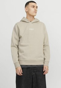 Beige pullover hoodie with a front kangaroo pocket, ribbed cuffs and hem, featuring white logo text on the chest, worn over a dark shirt.