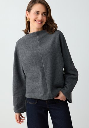 Donna sorridente con orecchini a cerchio in oro, una maglia grigia ampia a maniche lunghe e jeans blu scuro su uno sfondo chiaro.