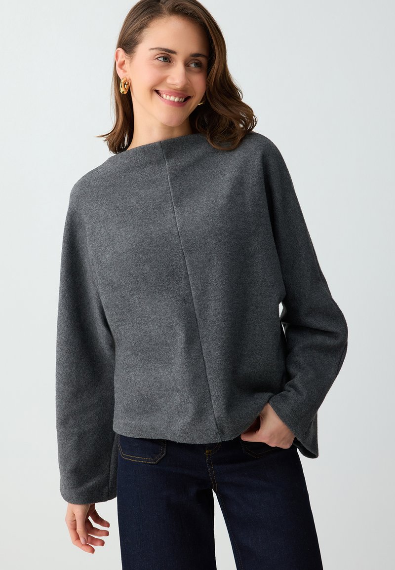 Femme souriante portant des boucles d'oreilles créoles en or, un haut gris ample à manches longues et un jean bleu foncé sur un fond clair.