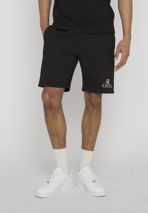 Homme portant un short noir avec un petit chien et le texte « CHILL » sur la jambe gauche, chaussettes blanches et baskets blanches, debout, les mains détendues.