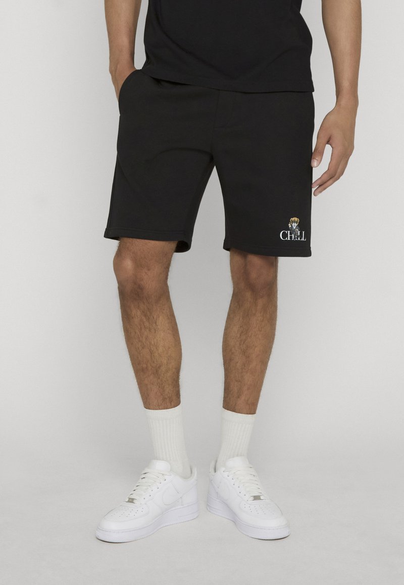 Homme portant un short noir avec un petit chien et le texte « CHILL » sur la jambe gauche, chaussettes blanches et baskets blanches, debout, les mains détendues.