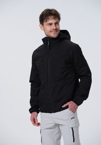 Schwarze wasserdichte Jacke mit Reißverschluss, elastischen Ärmelbündchen und zwei Brusttaschen, getragen mit hellen Cargo-Hosen.