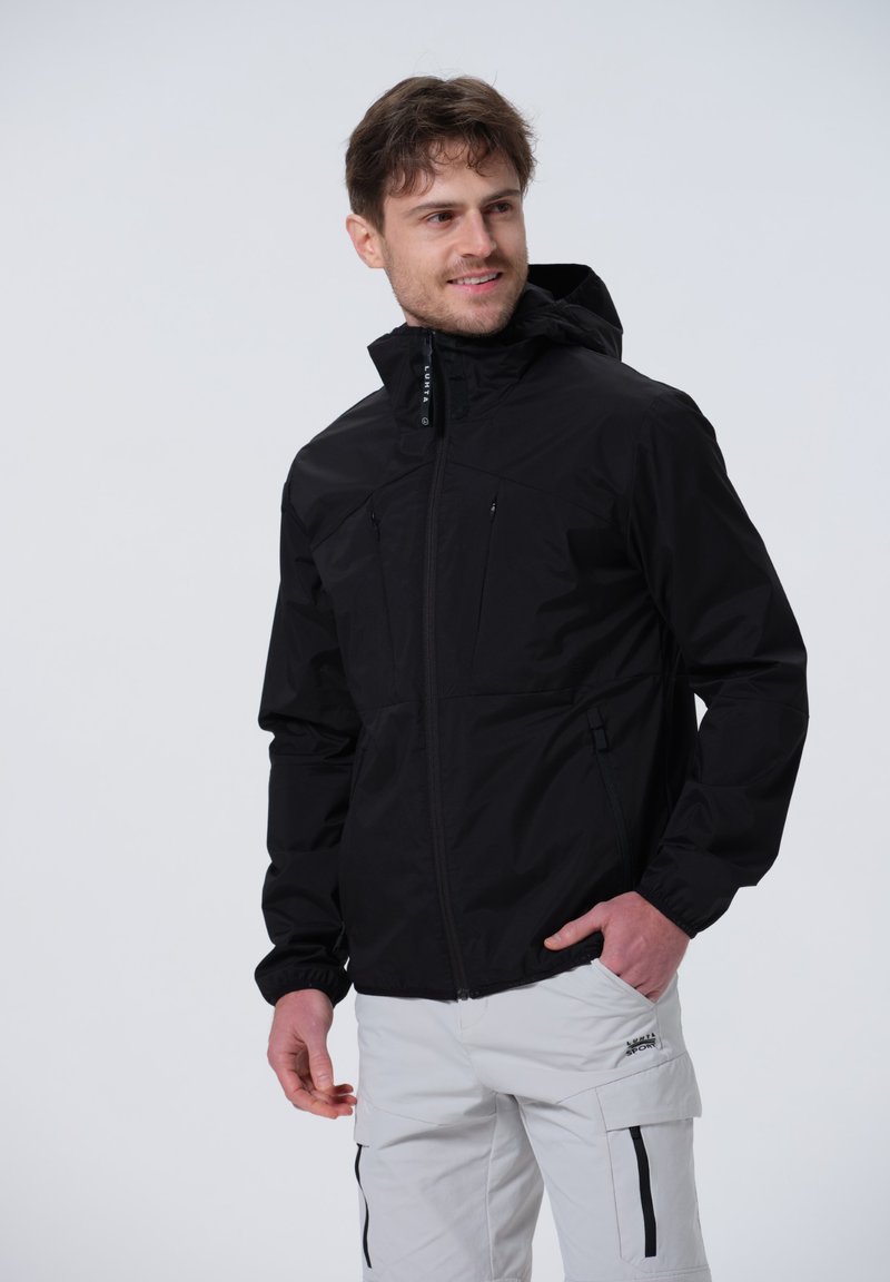 Schwarze wasserdichte Jacke mit Reißverschluss, elastischen Ärmelbündchen und zwei Brusttaschen, getragen mit hellen Cargo-Hosen.