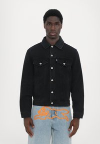 Veste en denim noir avec un col pointu, des poches avant et des boutons en argent. Associée à un jean bleu clair avec un design graphique orange.