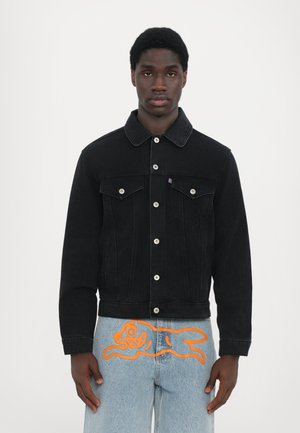 Zwarte spijkerjas met een spitse kraag, voorzakken en zilveren knopen. Gecombineerd met lichtblauwe jeans met een oranje grafisch ontwerp.