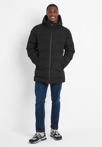 Black puffer jacket with a hood, zippered front, quilted design, and side pockets. Worn with blue jeans and sporty black and white shoes.