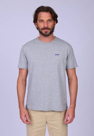 Homme barbu portant un t-shirt gris clair avec un petit logo bleu sur la poitrine et un pantalon beige, debout devant un fond clair uni.
