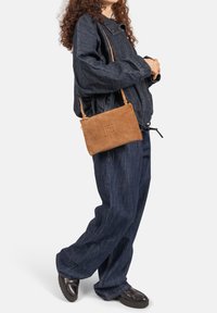 Bruine suède crossbodytas met een vlakke rechthoekige vorm, geperste details en een dun verstelbaar bandje, gedragen bij een donker denim outfit.
