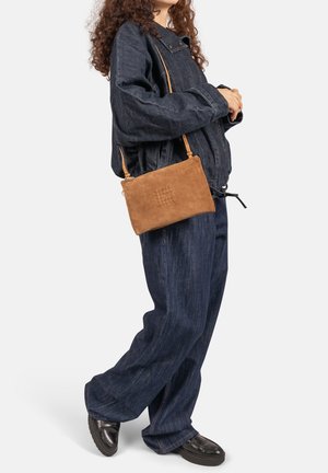 Bruine suède crossbodytas met een vlakke rechthoekige vorm, geperste details en een dun verstelbaar bandje, gedragen bij een donker denim outfit.