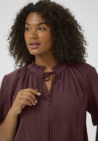 Burgundy crinkled blouse with a ruffled collar, tie detail, and button placket. Short sleeves and textured fabric create a relaxed fit.