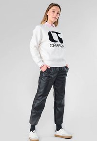 Sudadera blanca con logo negro, superpuesta a una camisa de color rosa claro con cuello. Pantalones de cuero sintético negros, zapatillas blancas y calcetines oscuros.