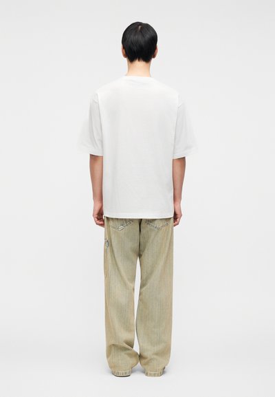 Λευκό βαμβακερό T-shirt με άνετη εφαρμογή, συνδυασμένο με μπεζ τζιν παντελόνι φαρδιάς γραμμής, που έχει πλαϊνή τσέπη με φερμουάρ και διακριτικές ραφές.