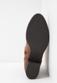 Sohle eines braunen Lederdressschuhs, der an einer weißen Ecke lehnt, mit detaillierter schwarzer Gummisohle und -absatz.