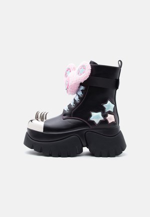 Černá platformová bota se stříbrnou špičkou ve tvaru drápu, růžovými chlupatými klipy na uších s modrými hvězdami a modrými a růžovými hvězdnými nášivkami na boku.