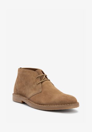 Suède chukka-laars in lichtbruin met een ronde neus, minimale vetersluiting en tonale stiksels. Voorzien van een platte rubberen zool voor comfort.