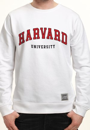 Person iført en hvid sweatshirt med "HARVARD" i store røde bogstaver og "UNIVERSITY" i mindre sorte bogstaver nedenunder.