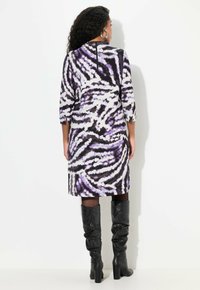 Lila und schwarz gemustertes Kleid mit Dreiviertelärmeln, knielanger Schnitt und einem rückseitigen Reißverschluss. Getragen mit schulterhohen schwarzen Stiefeln.