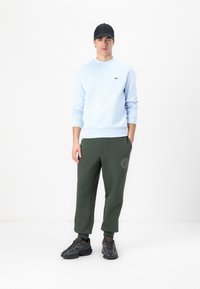 Lyseblå hettegenser med ribbestrikkede mansjetter og brodert logo, kombinert med mørkegrønne joggebukser med en sidelogo-detalj, og fullført med svarte joggesko.