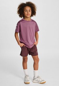 Koszulka sportowa z krótkim rękawem w kolorze liliowym, z akcentami na ramionach, w połączeniu z ciemnobrązowymi szortami. Całość dopełniają białe buty sportowe i skarpetki do kostek.