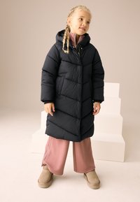 Manteau matelassé noir avec capuche, présentant une texture matelassée et des coutures verticales. Associé à un pantalon rose à jambes larges et à des bottes beige.
