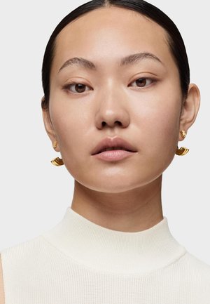 Pendientes de declaración dorados con un diseño ondulado, que presentan acentos texturizados. La modelo lleva una blusa blanca de cuello alto y con relieve.