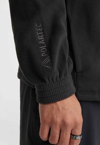 Musta fliisjakker, millel on venivad mansetid, õlavarre kohal on õrn reljeefne "Polartec" logo ning kangas on sileda tekstuuriga.