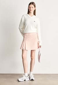 Crème V-halstrui, roze gestreepte skort met plooien en witte sneakers. Klein groen logo op de skort. Houdt een roze handtas vast.
