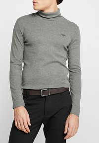 Maglione tunica grigio in un tessuto morbido con maniche lunghe, caratterizzato da un piccolo logo sul petto, abbinato a pantaloni neri e una cintura marrone.