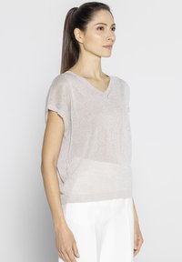 Jasno beżowa bluzka z krótkim rękawem i dekoltem w serek, uszyta z miękkiego, lekko błyszczącego materiału, o luźnym kroju, zestawiona z białymi spodniami o szerokich nogawkach.