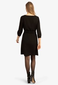Schwarzes Kleid mit Dreiviertelärmeln, gerafftem Taillenteil und rundem Ausschnitt. Getragen mit schwarzen High-Heel-Stiefeln und transparenten Strumpfhosen.