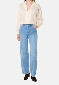Camicia button-up beige chiaro con colletto, abbinata a jeans dritti a vita alta blu chiaro e stivali alla caviglia scuri.
