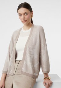 Jasno beżowy kardigan z wzorzystym splotem, szerokimi rękawami i otwartym przodem, narzucony na biały top z prążkowanego materiału oraz beżowe spodnie.