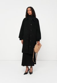 Cappotto in misto lana nero con colletto e sei bottoni, abbinato a una gonna plissettata nera. Tacchi neri con cinturini e borsa a spalla beige.