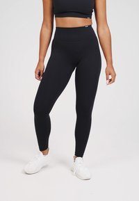 Leggings negros de cintura alta hechos de una tela elástica. Textura suave con un diseño ajustado, combinados con un sencillo top corto negro y zapatillas blancas.