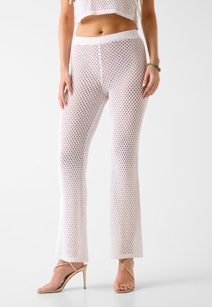 Kobieta ubrana w białe siateczkowe spodnie i dopasowany krótki top, nosząca złote sandały na wysokim obcasie, stojąca na jednolitym tle.