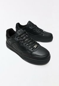 Paire de baskets basses noires en cuir texturé, lacets noirs et semelles épaisses en caoutchouc sur fond blanc.