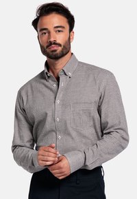 Man met een baard in een gestreepte button-up shirt die zijn manchet aanpast, staand tegen een effen witte achtergrond.