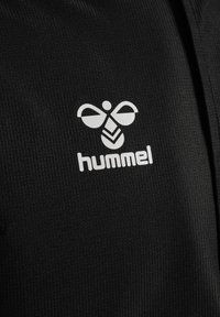 Białe stylizowane logo pszczoły i tekst „hummel” nadrukowane na czarnej materiałowej tkaninie o fakturze, prawdopodobnie materiał sportowy.