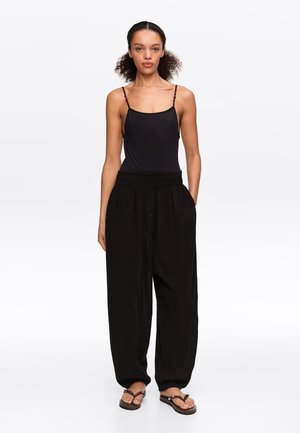 Kobieta stoi zwrócona przodem, ubrana w czarny bezrękawnik, luźne czarne spodnie zapinane na guziki z kieszeniami oraz czarne sandały.