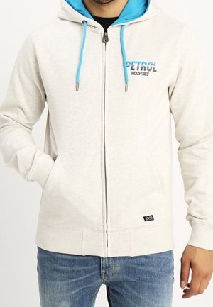 Homme portant un sweat à capuche gris clair zippé avec des cordons bleus et le logo "Petrol Industries" sur la poitrine, assorti à un jean bleu.