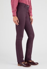 Pantalones slim-fit burdeos hechos de una tela suave, con un diseño clásico de cinco bolsillos y un cierre frontal con botón, combinados con zapatos brillantes.