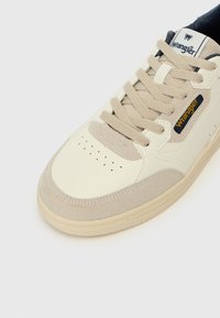 Baskets crème et beige avec une tige en cuir et en suède, bout arrondi, lacets épais, et logo Wrangler sur le côté et la languette.