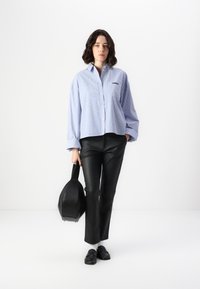 Blå og hvit stripete oversized skjorte med knapper, sorte skinnbukser, holder en svart rund håndveske, svarte loafers med hvite sokker.