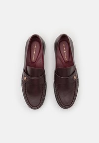 Chaussures slip-on en cuir bordeaux au toucher lisse, ornées d'une sangle décorative et d'un détail logo en or sur le dessus. Semelle en caoutchouc noir.
