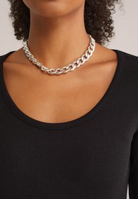 Collar de cadena de plata con grandes eslabones entrelazados y acabado brillante. Lleva puesto sobre una blusa negra de canalé con escote en forma de barco. Vista en primer plano.