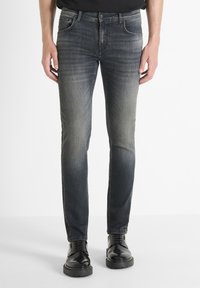 Mršave traperice tamnog pranja, s izblijedjelom gradijentnom bojom, pet džepova i žutim šavovima. Uparene s crnim cipelama na vezanje.