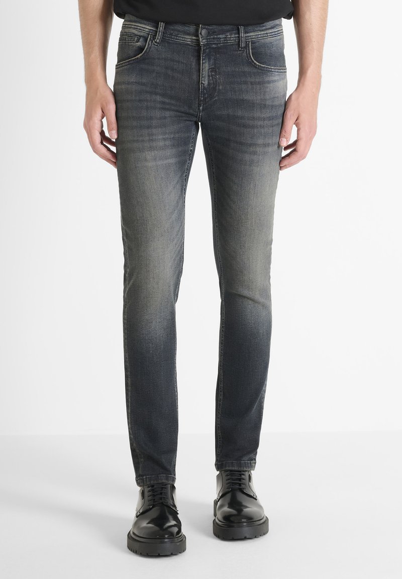 Mršave traperice tamnog pranja, s izblijedjelom gradijentnom bojom, pet džepova i žutim šavovima. Uparene s crnim cipelama na vezanje.