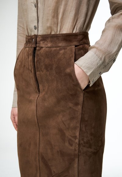 Personne portant une jupe en daim marron avec une fermeture éclair à l'avant et une chemise beige à boutons, main dans la poche de la jupe, gros plan sur la taille et le torse.