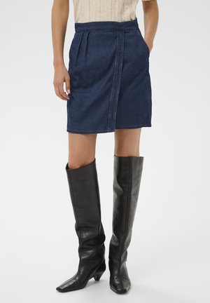 Frau, die einen blauen Jeansrock und schwarze kniehohe Lederstiefel trägt, steht mit einer Hand in der Tasche.