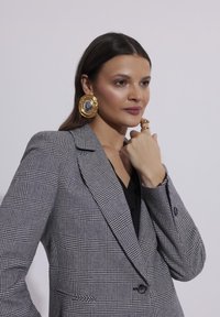 Szary w kratę marynarka o dopasowanym kroju, czarna bluzka z dekoltem w serek pod spodem oraz duże, złote okrągłe kolczyki. Gładki materiał z widoczną teksturą.